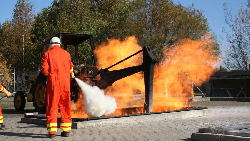 Das Foto zeigt eine Person im Schutzanzug, die ein Feuer in einer nachgestellten Baustellengrube löscht.